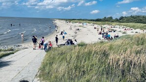 A beach with people walking and sitting, a concrete path, and grassy dunes. - Holiday Home Anje Comfortable Vacation Home (Krummhoern)