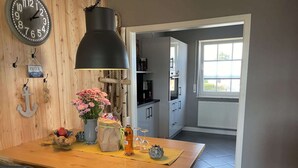 A dining area with a wooden table, chairs, a black pendant light, a clock, and a view into the kitchen. - Holiday Home Anje Comfortable Vacation Home (Krummhoern)