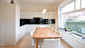 A modern kitchen with white cabinets, a wooden dining table, and a view of the neighborhood through large windows. - Apartment Near Ostend Beach (De Haan)