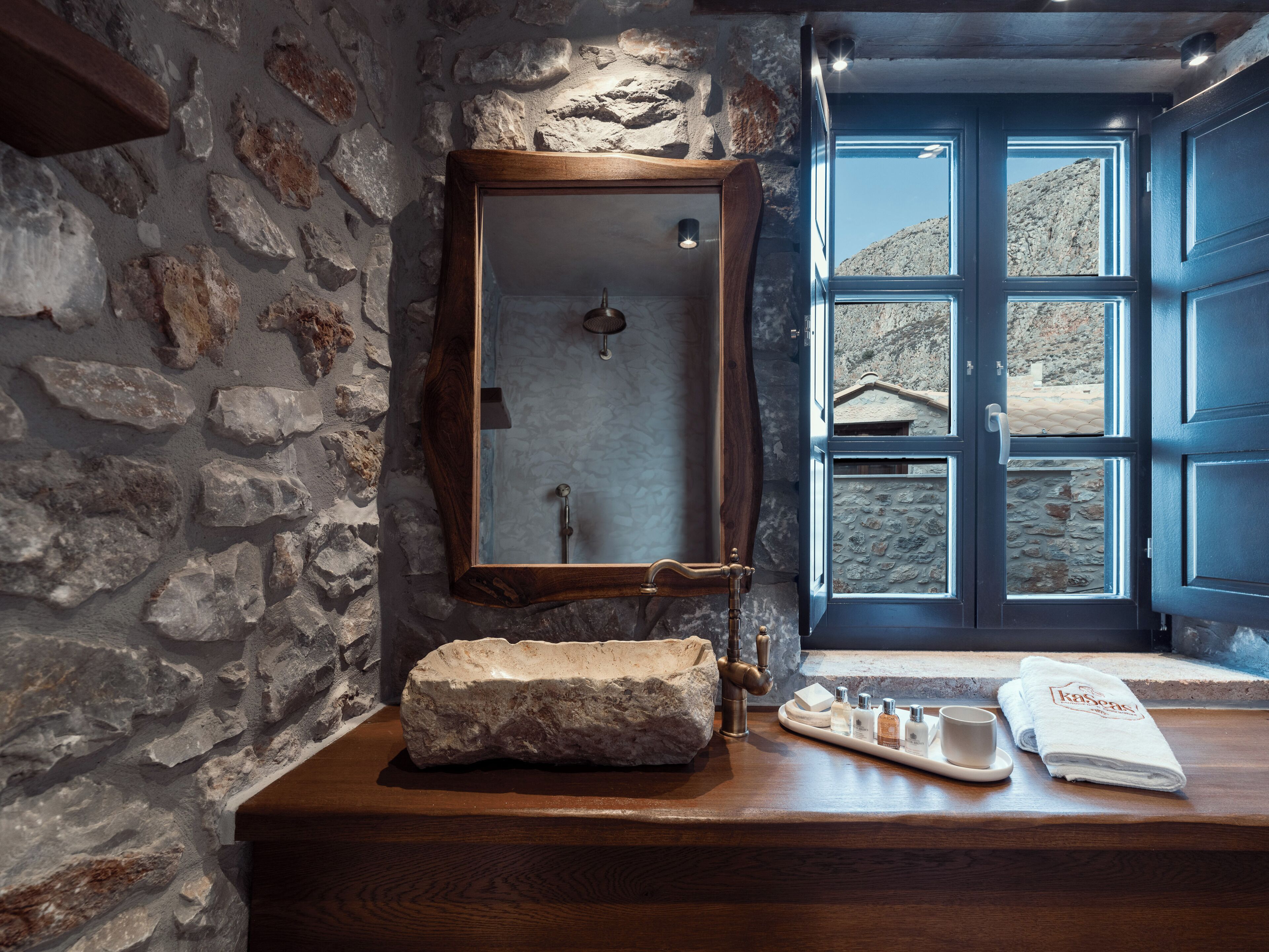 Een badkamer met een stenen muur, een houten wastafel, een grote spiegel, een douche en uitzicht buiten door een blauwe deur.