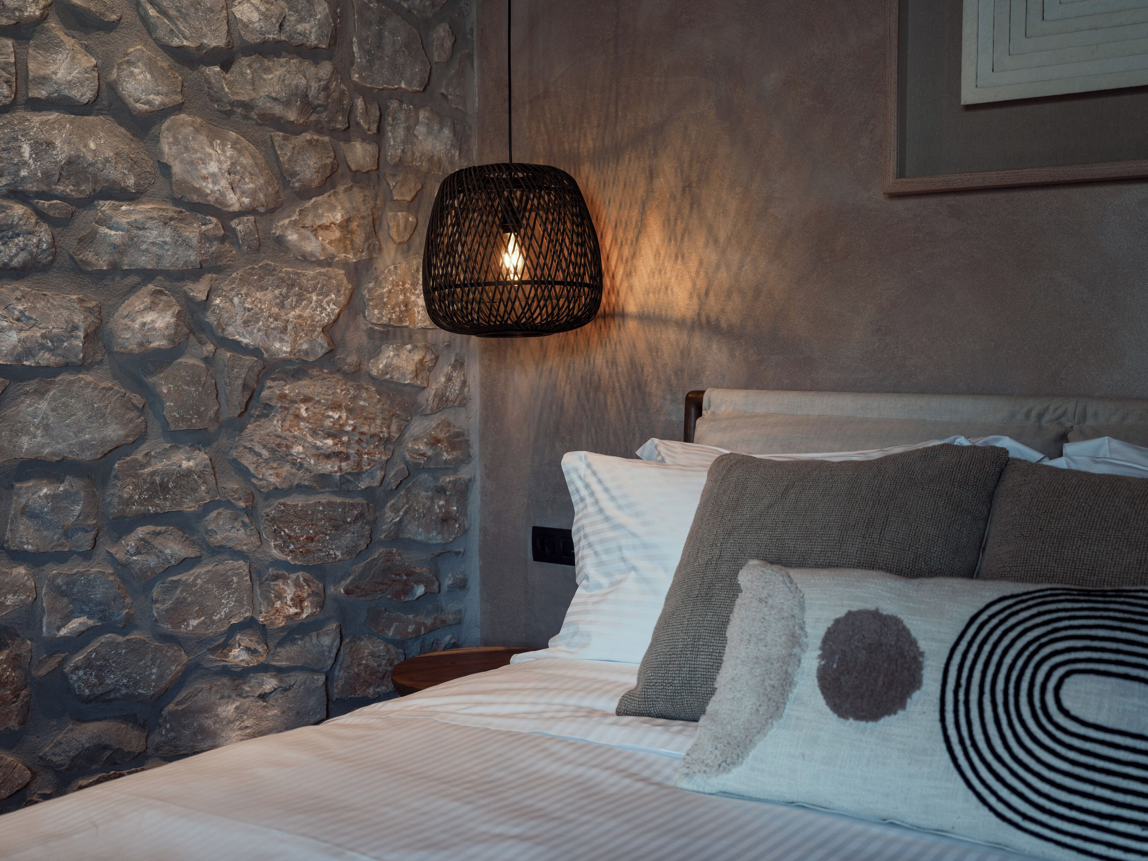 A bedroom with a stone wall, a hanging light fixture, a bed with white bedding and patterned pillows, and a wooden nightstand.