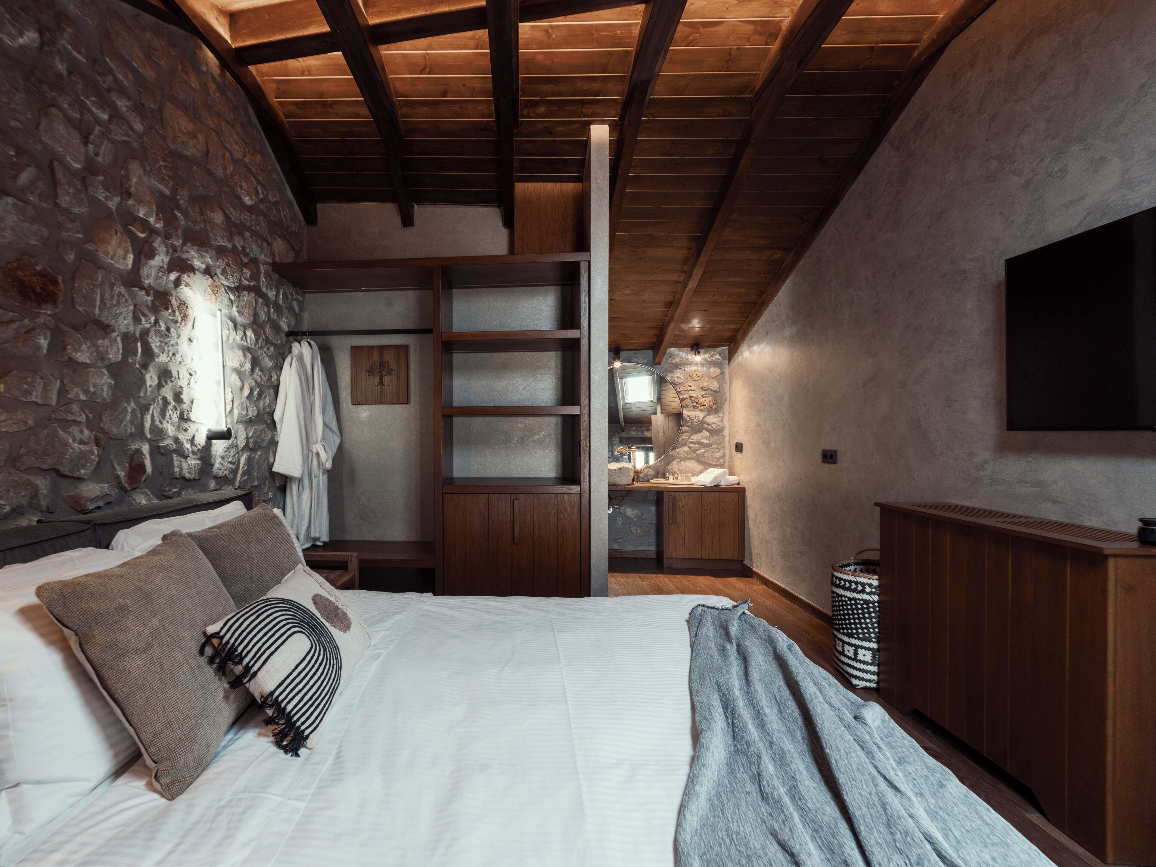 A bedroom with a stone wall, wooden beams, a bed with pillows, a wardrobe, a mirror, and a television.