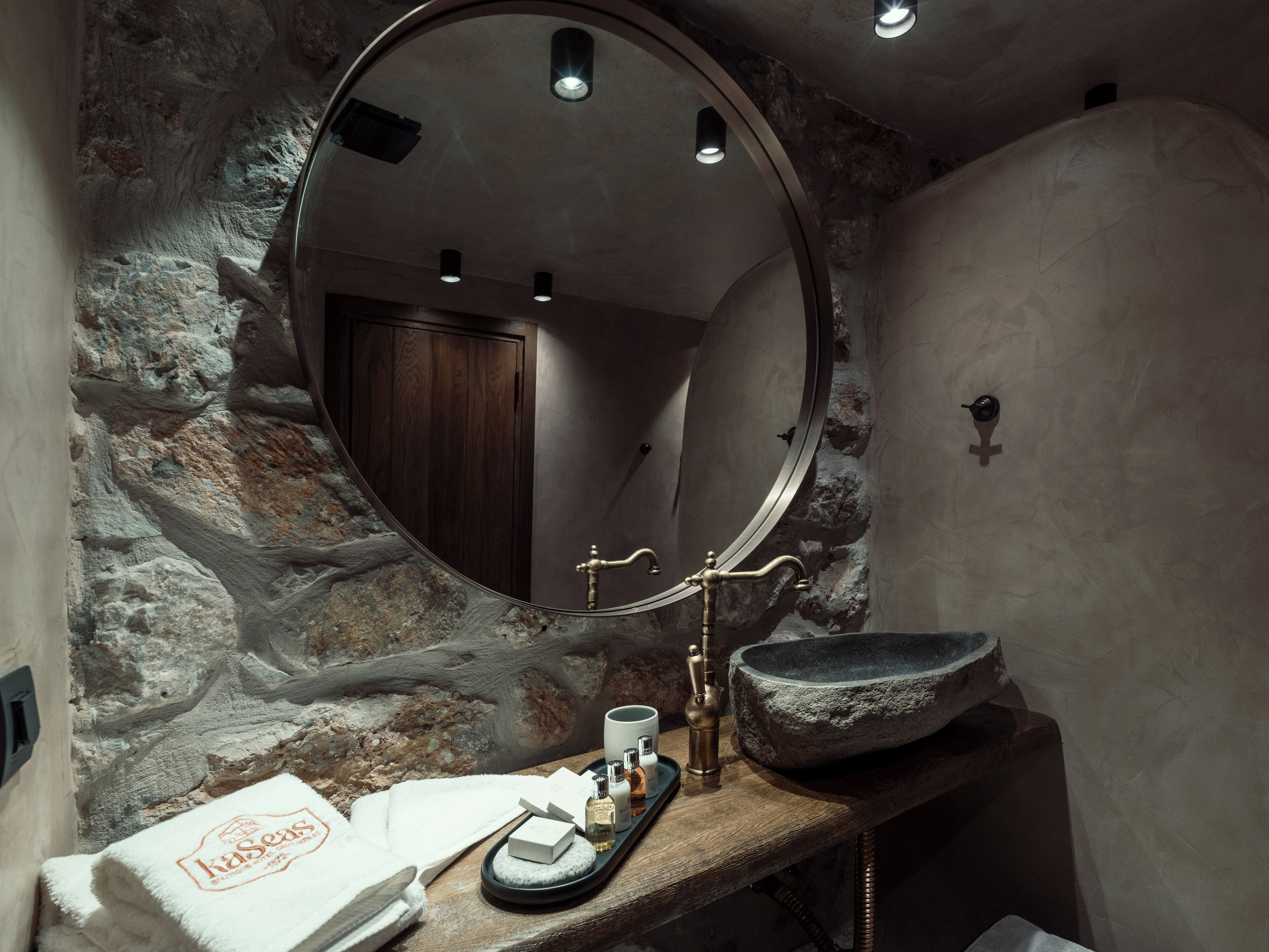 A bathroom with a stone wall, a round mirror, a wooden door, a sink with a stone basin, and toiletries on a tray.