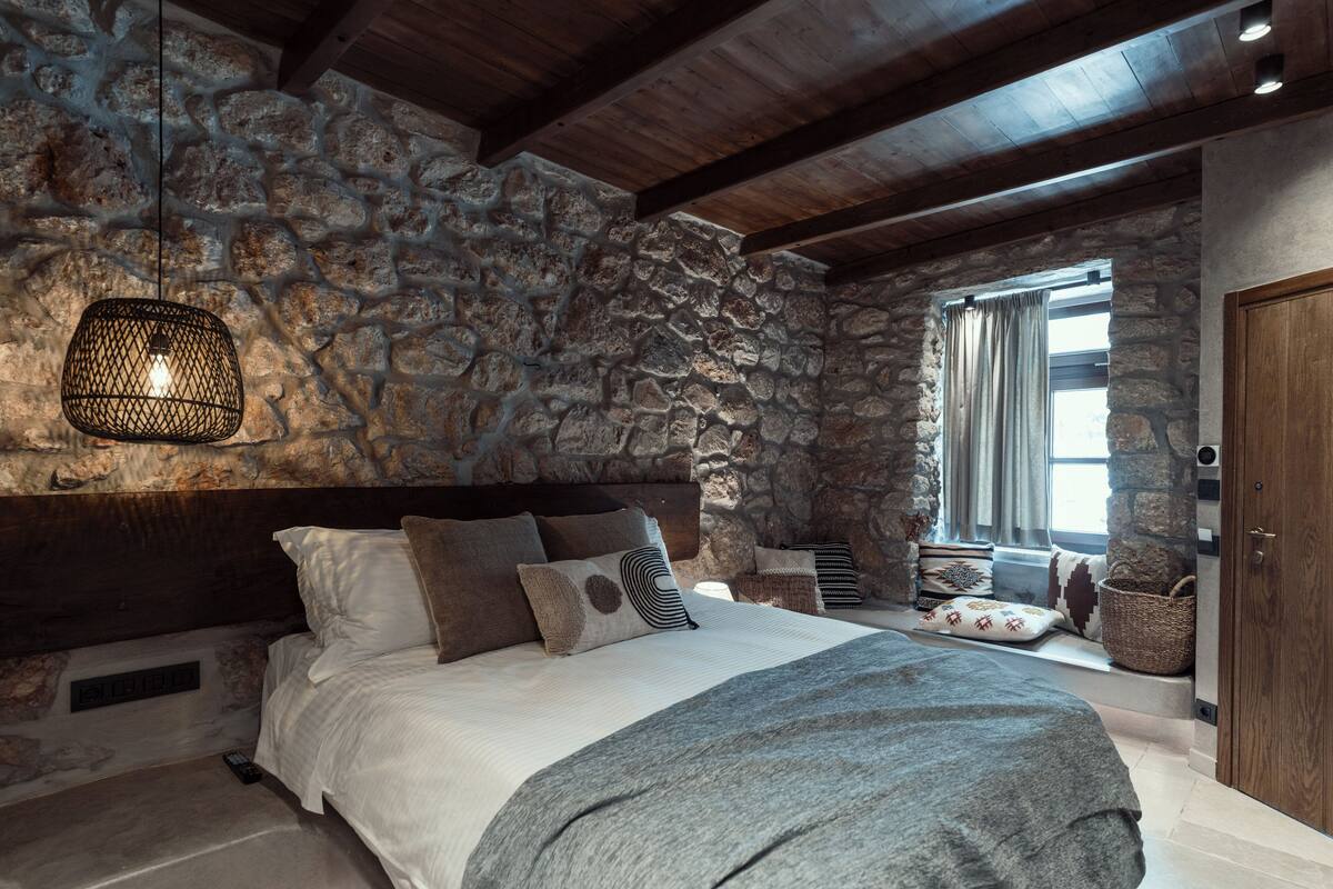 Een slaapkamer met een stenen muur, houten balken, een bed met kussens, een hanglamp en een raam met gordijnen.
