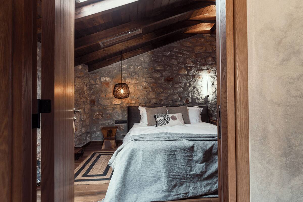 Een slaapkamer met een stenen muur, houten balken in het plafond, een bed met een grijze deken en een hangende lamp.