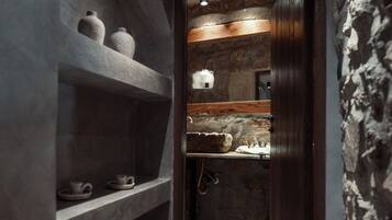 A narrow hallway with stone walls and wooden shelves. There are three light fixtures on the ceiling, and a wooden door at the end.