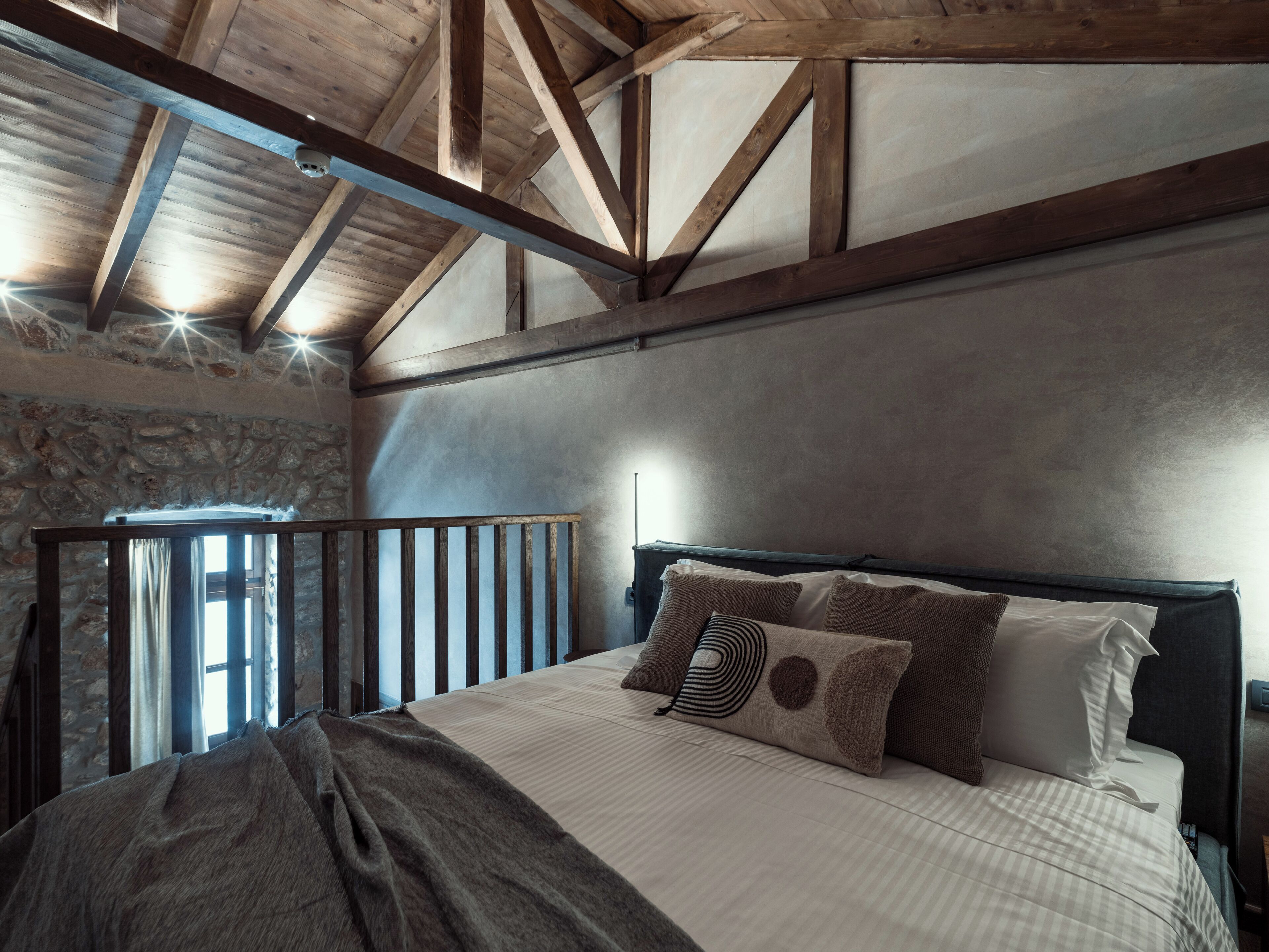Un dormitorio con techo de madera, pared de piedra y una cama con muchos almohadones.