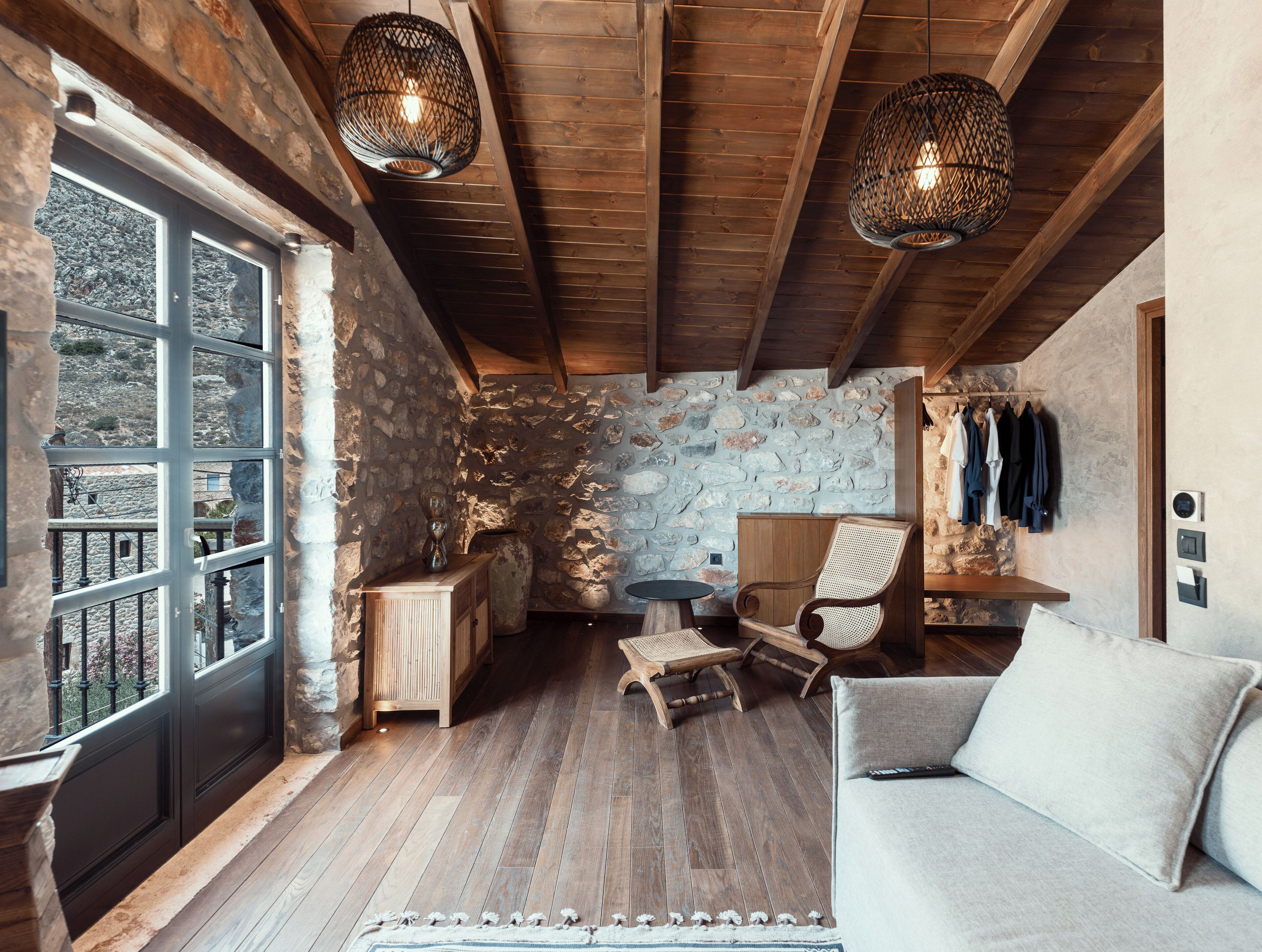 A rustic interior with stone walls, wooden beams, a stone fireplace, a wooden bench, a woven chair, and a sofa.