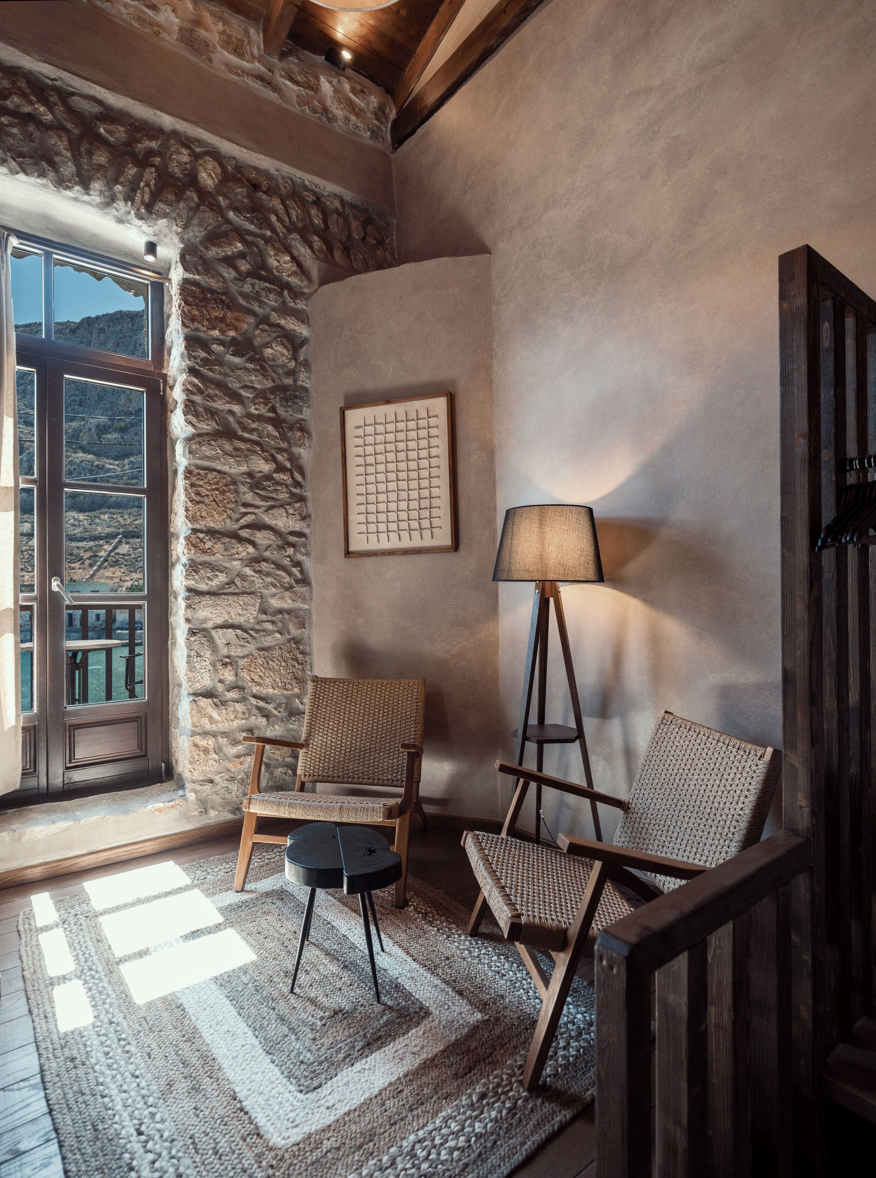 Una habitación acogedora con paredes de piedra, un ventanal grande, dos sillones de mimbre, una lámpara de pie y una obra de arte enmarcada.