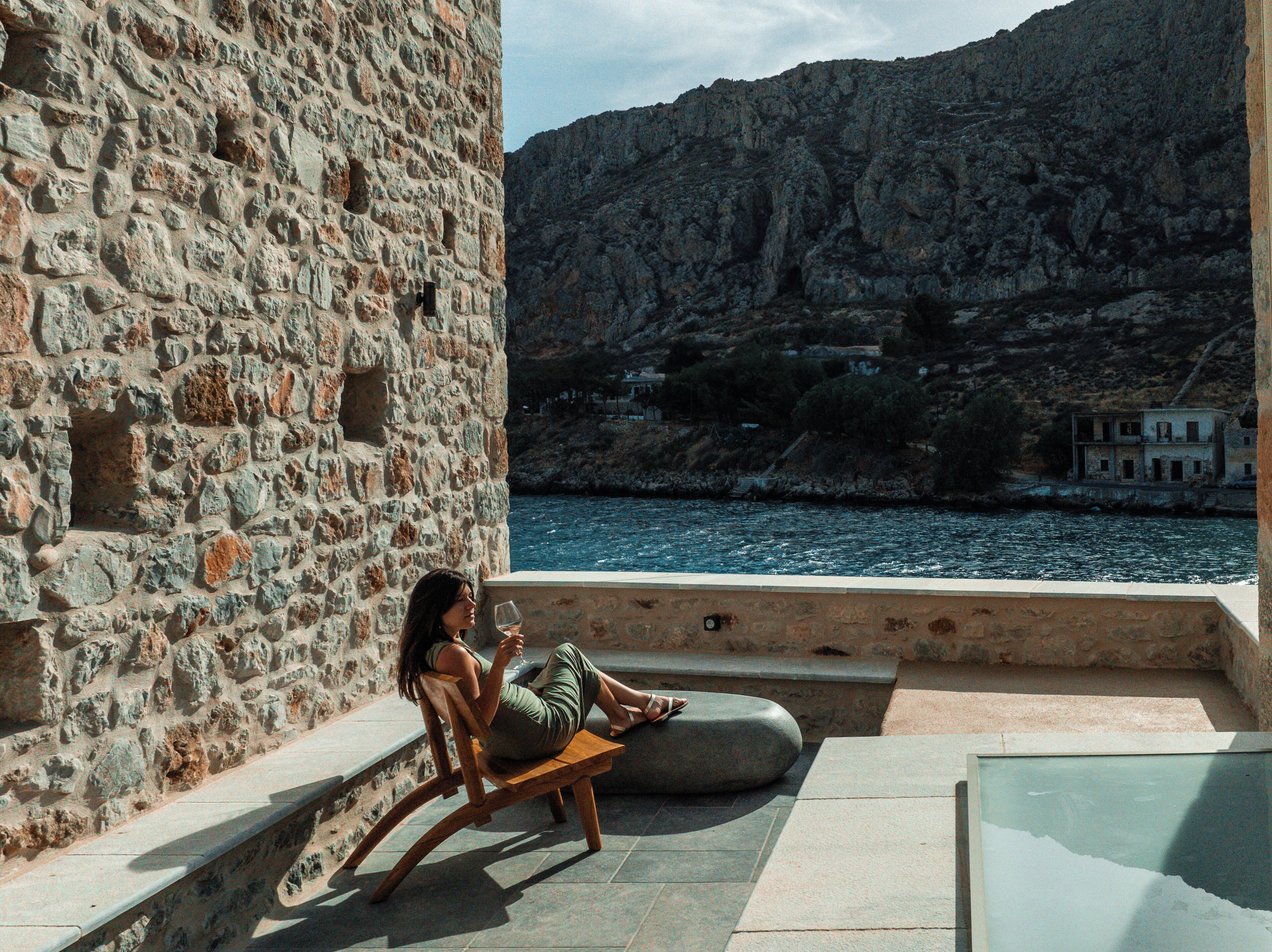 Un mur de pierres sur lequel est assise une femme, sur un banc, d’où elle domine un plan d’eau et un paysage montagneux.