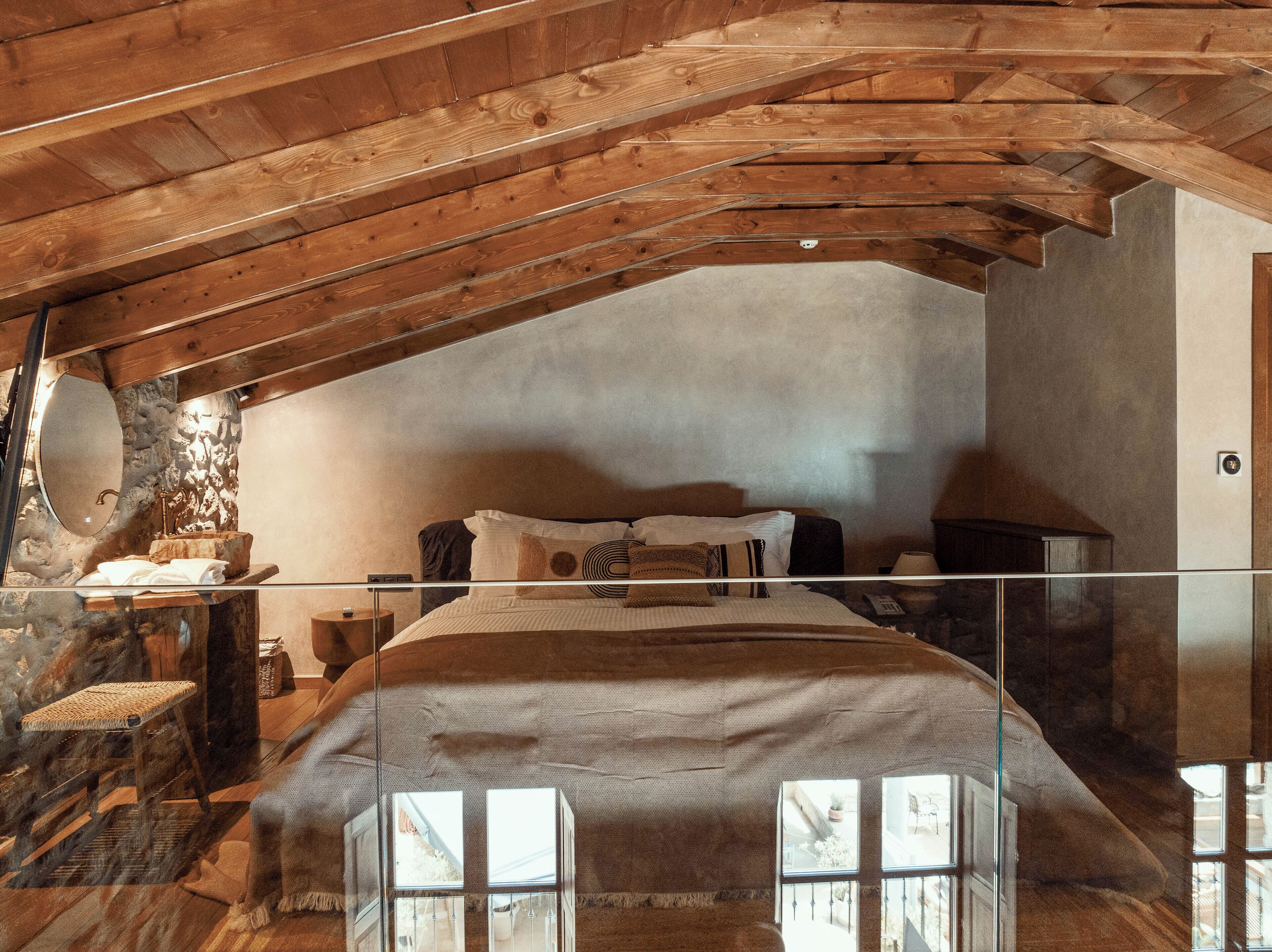 Un dormitorio rústico con techo inclinado, una cama con cabecera de madera, pared de piedra y una mesita con lámpara.