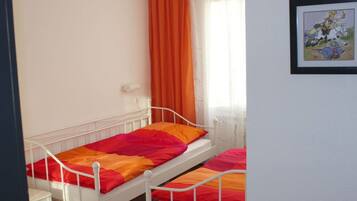Ein Zimmer mit zwei Einzelbetten, einem Fenster mit orangefarbenen Vorhängen und einem gerahmten Bild an der Wand.