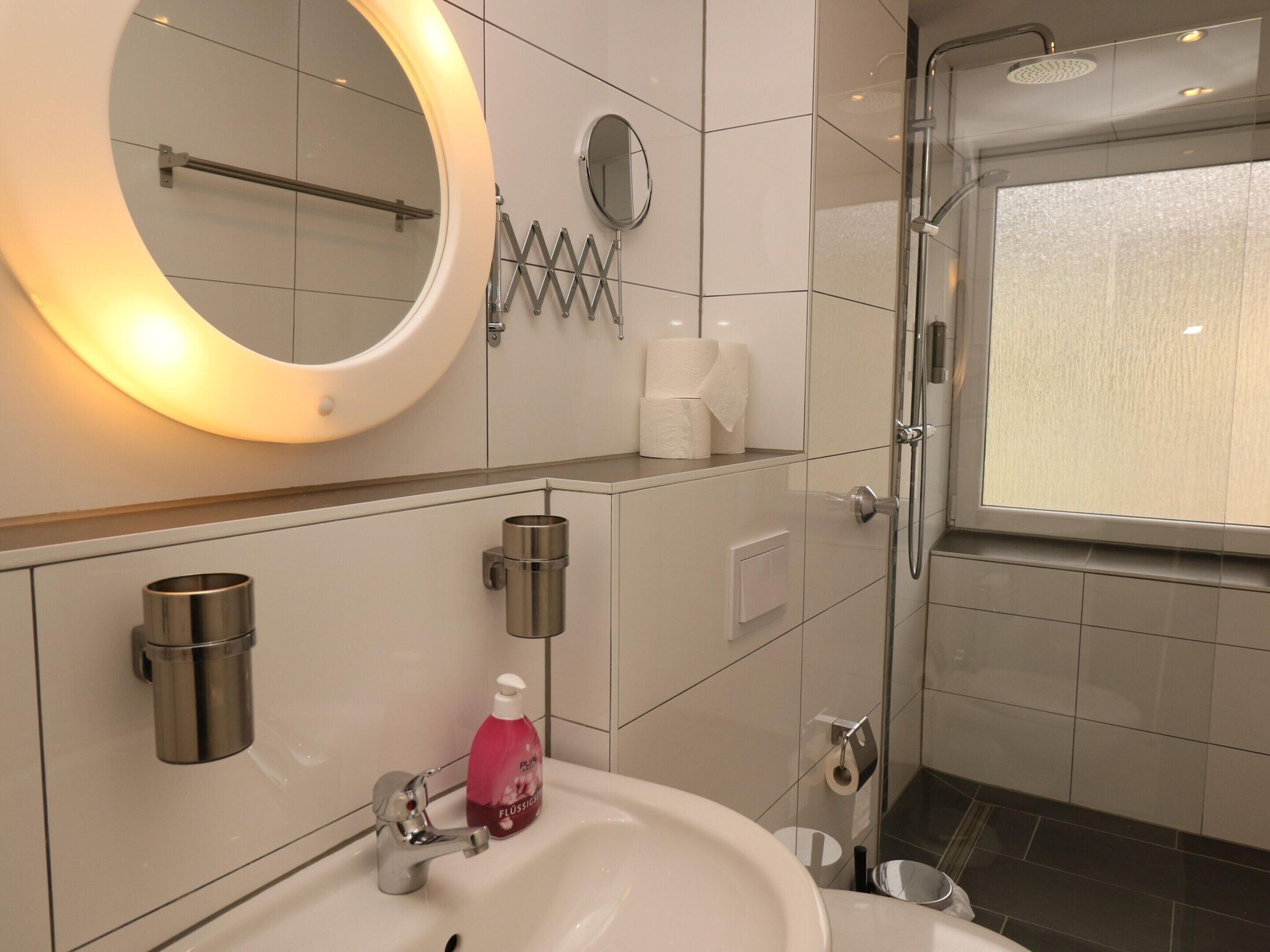 A modern bathroom with a round mirror, a sink, a shower, and a toilet paper holder.
