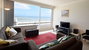 Ein modernes Hotelzimmer mit großem Fenster und Meerblick, einem Flachbildfernseher, einer Couch und einem kleinen Tisch mit Lautsprechern.