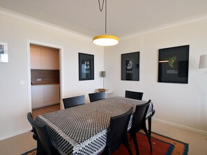 A dining area with a round table, black chairs, and a patterned tablecloth. There are framed artworks on the walls and a modern pendant light above the table. - Coastal Comfort in De Haan (De Haan)