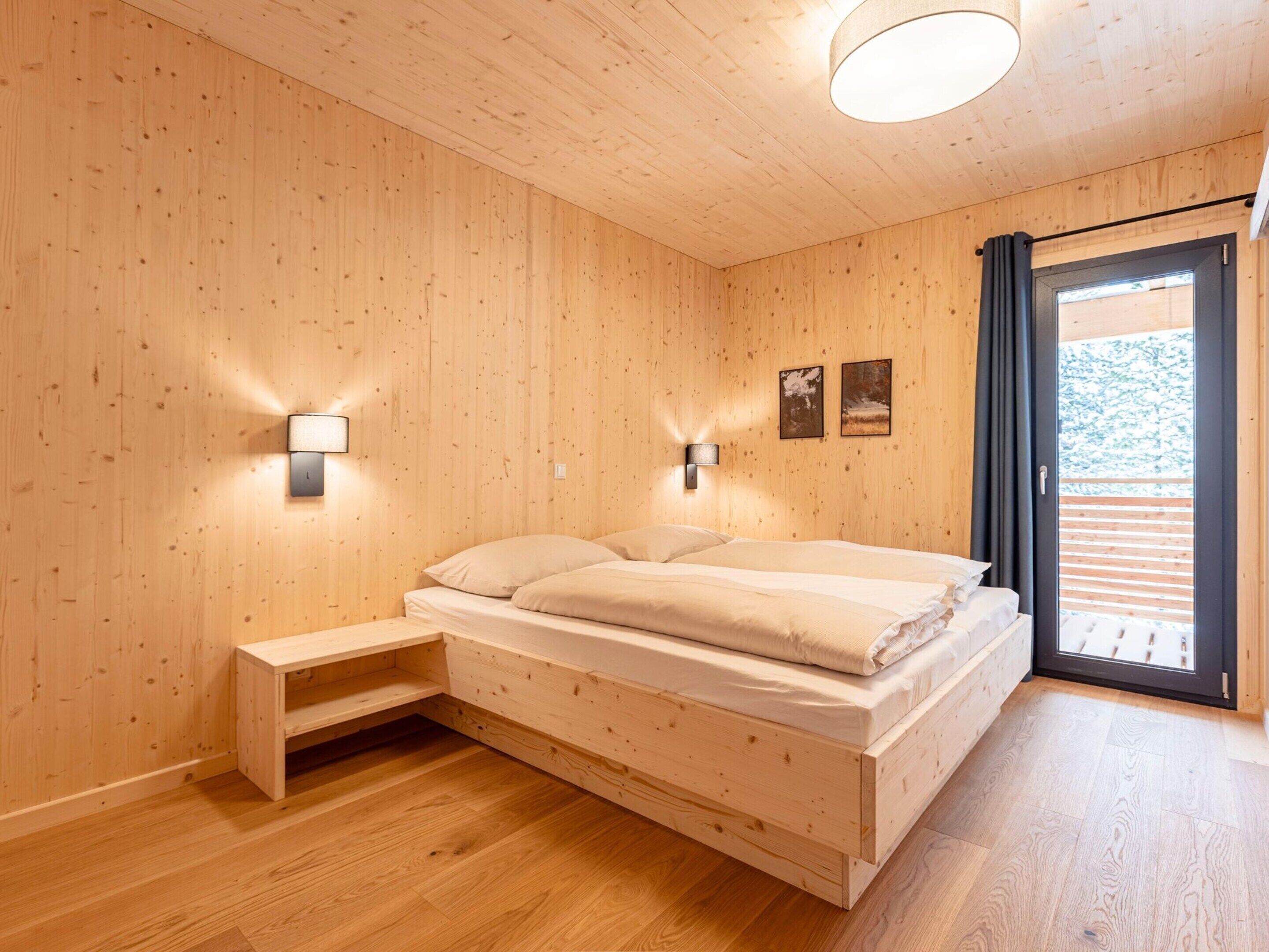 A wooden-paneled room with a bed, a bedside table, two wall-mounted lights, and a window with curtains.