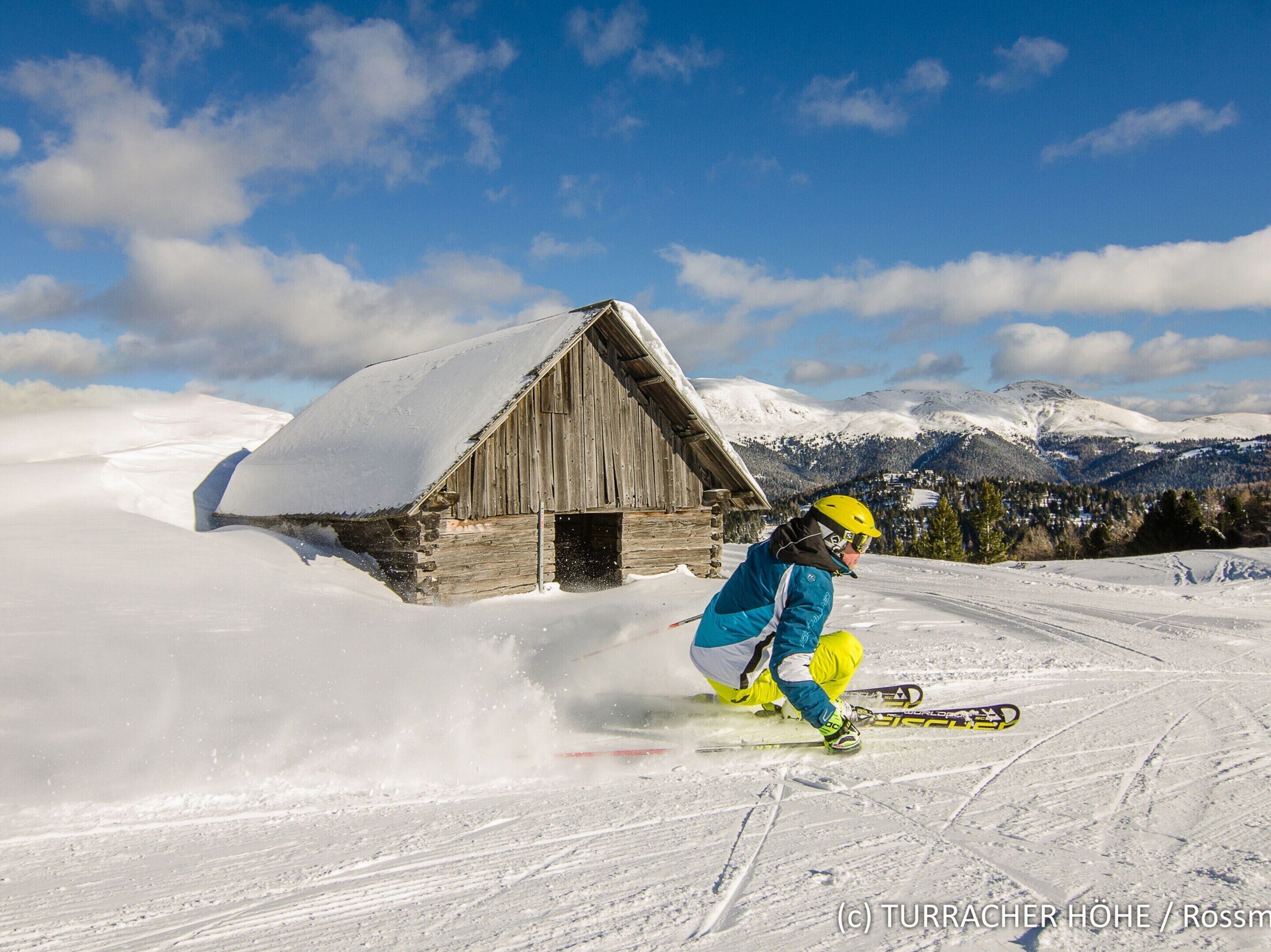 Eine Holzhütte mit schneebedecktem Dach, umgeben von verschneiter Landschaft und Bergen im Hintergrund.