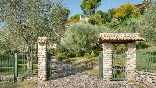 A stone gate with a tiled roof structure, surrounded by greenery and a fence. - Modern Holiday Home in Marciaga With Garden (Costermano sul Garda)