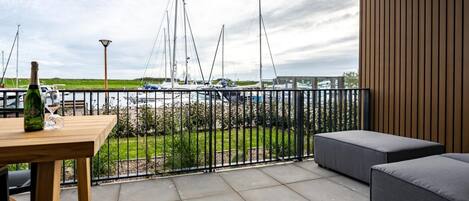 A balcony with a view of boats, a wooden table with a bottle and glass, and a grey sofa.