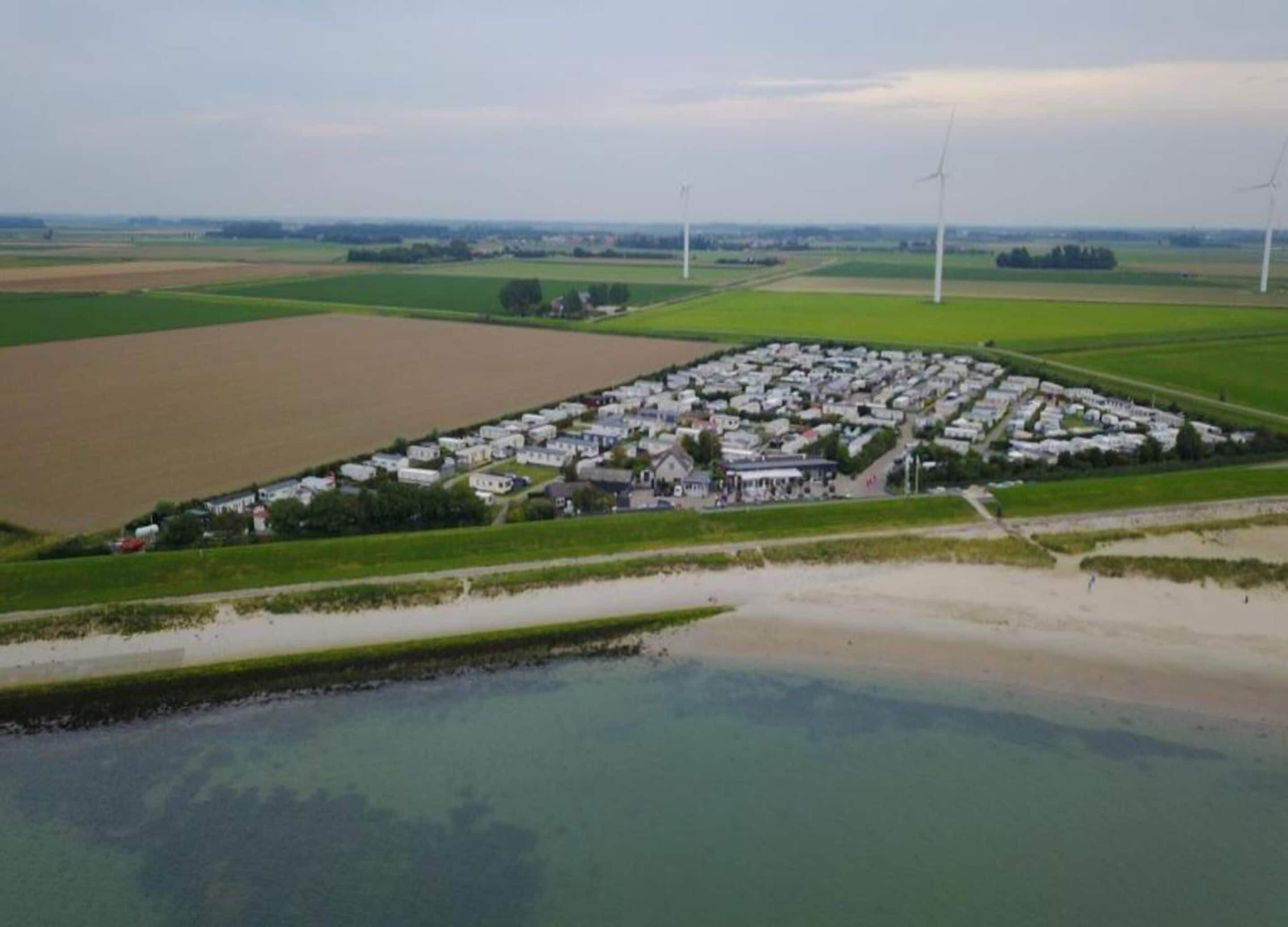 A large, rectangular campground with numerous RVs and trailers, surrounded by fields and wind turbines in the background.