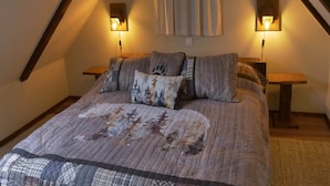 Une chambre à coucher accueillante avec un plafond en pente, un lit recouvert d’une courtepointe à motifs, deux lampes de chevet et une fenêtre avec des rideaux.