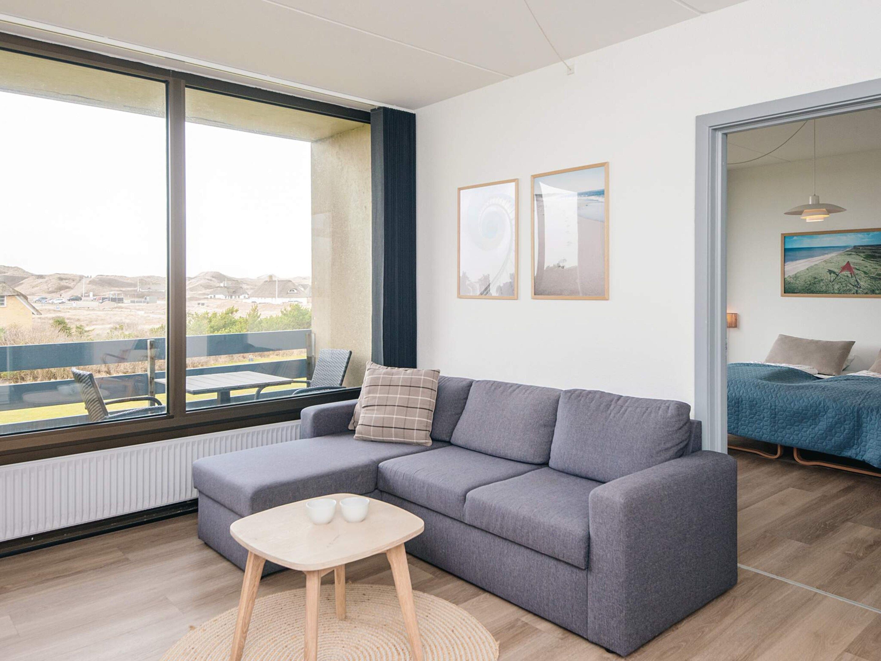 Una sala moderna con un sofá gris, una pequeña mesa de madera con dos tazas, un ventanal con vista a un paisaje y una puerta que da a un dormitorio.