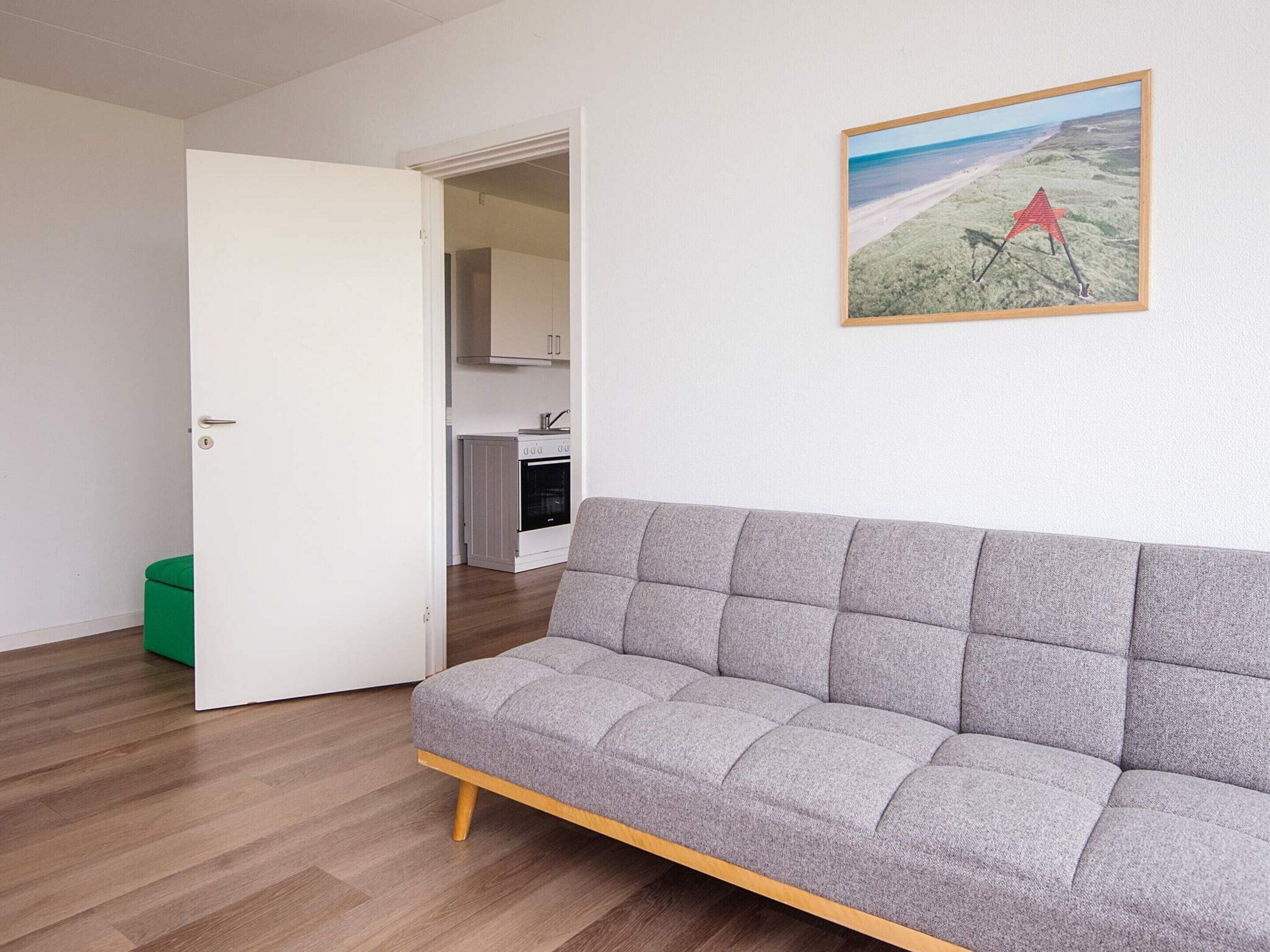 Et værelse med en grå sofa, en grøn skraldespand, en hvid dør og et køkken i baggrunden.