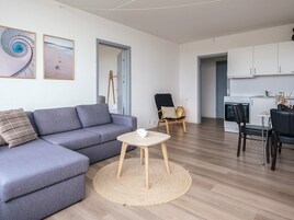 Una sala moderna con un sofá gris, una mesita redonda y una silla. Hay una zona de cocina con armarios blancos y una estufa.