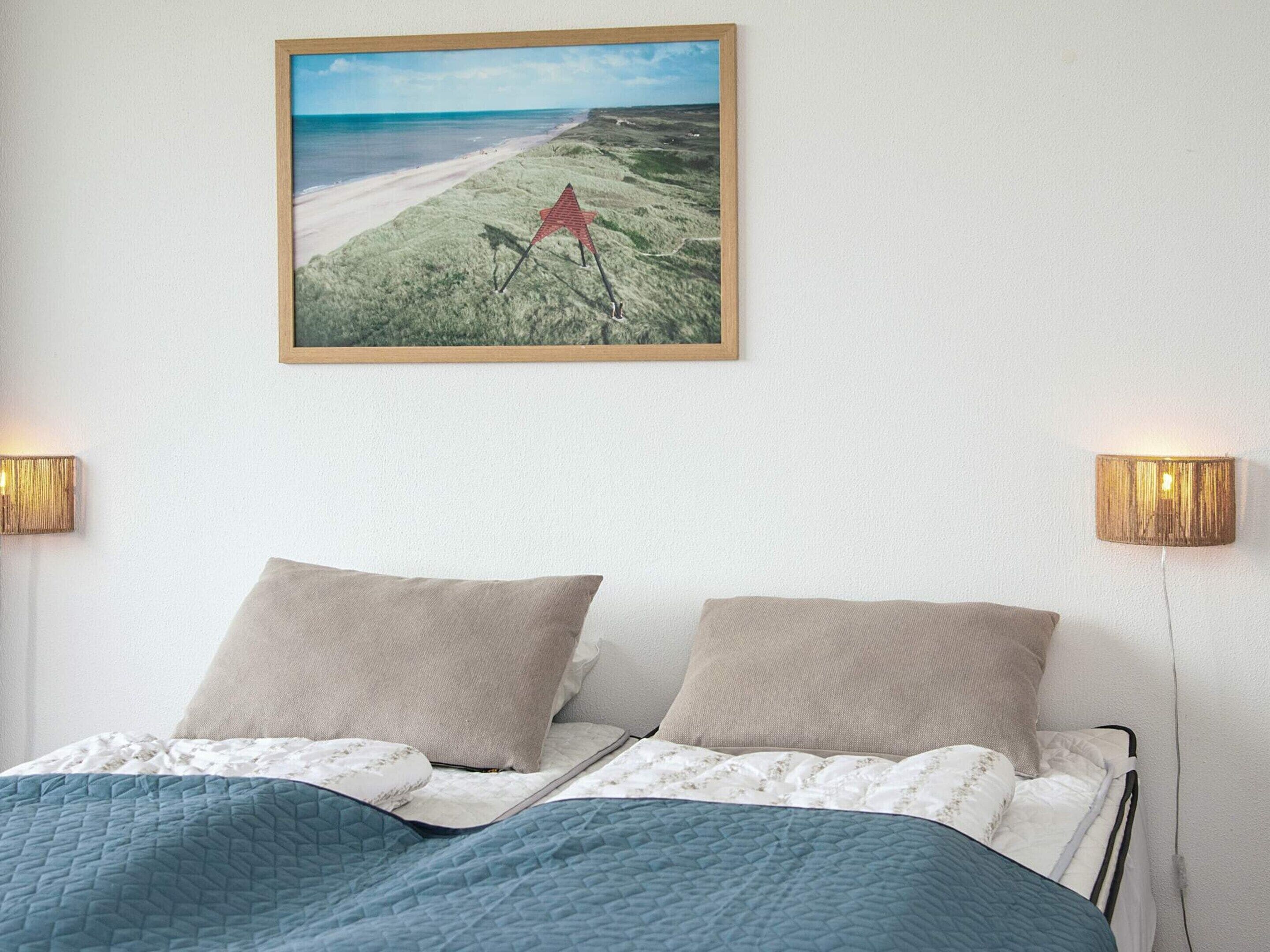 A bedroom with a bed, two pillows, a quilted blanket, a framed picture of a beach, and two wall-mounted lamps.