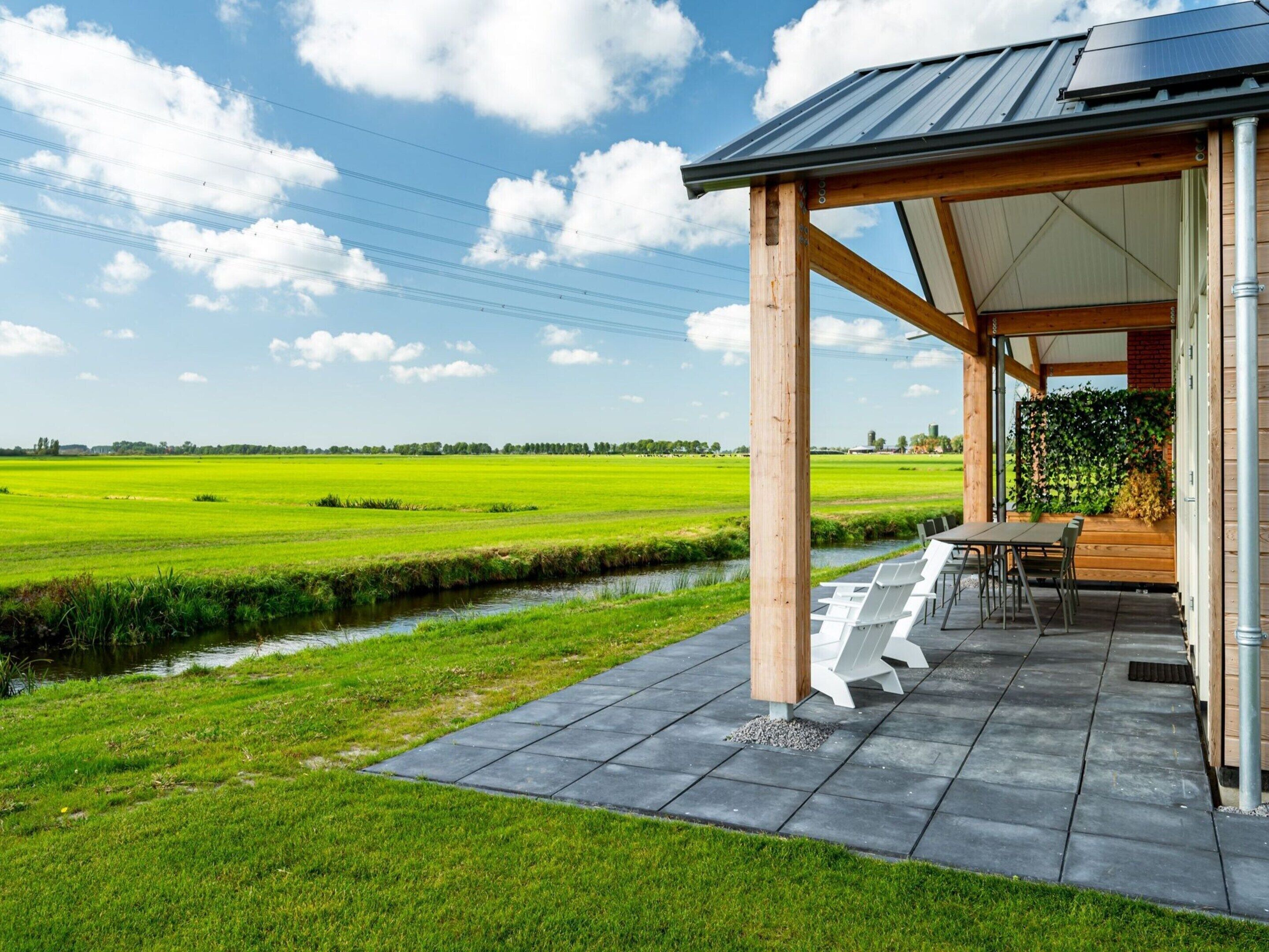 A covered outdoor seating area with a view of a green field and a small water channel.