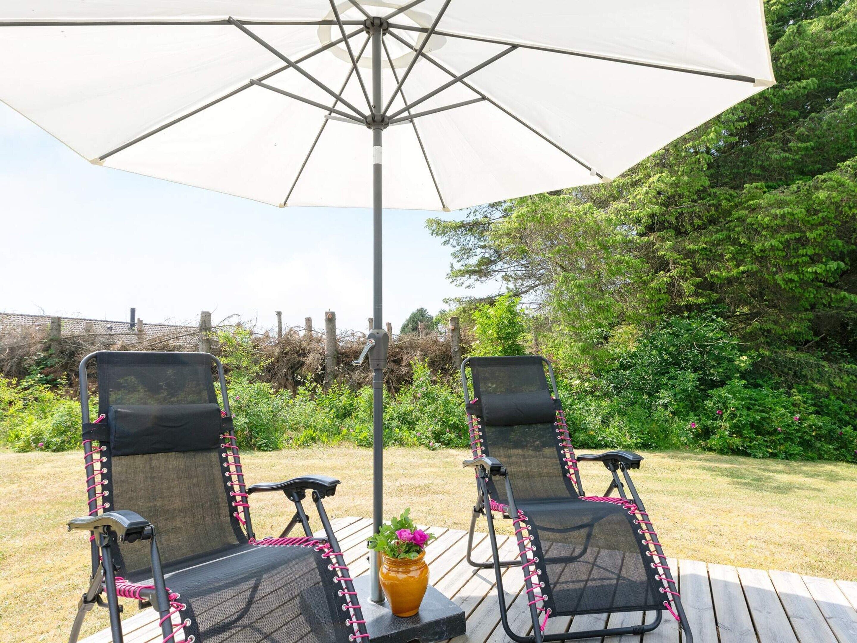 Two outdoor lounge chairs under a large umbrella, with a wooden deck and a garden in the background.