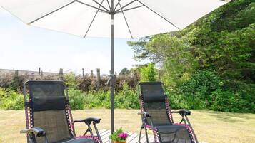 Two outdoor lounge chairs under a large umbrella, with a wooden deck and a garden in the background.