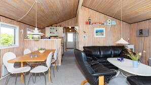 A modern cabin interior with a wooden ceiling, a dining area with a wooden table and white chairs, a black leather sofa, and a small white table with a vase. - 4 Person Holiday Home in Hjorring (Hjørring)