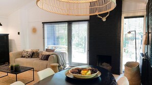 Uma sala de estar moderna com sofá, mesa de jantar com uma tigela decorativa, lareira e grandes janelas com persianas.