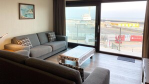 A modern living room with a grey sofa, a coffee table, and a large window offering a view of an industrial area. - Apartment in Nieuwpoort Near the Beach (Nieuwpoort)