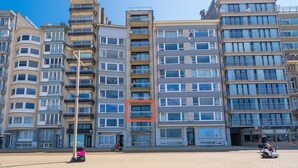 A multi-story residential building with balconies, a paved plaza, and a clear sky.
