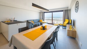 A modern hotel room with a dining area, a sofa set, and a view of the beach.