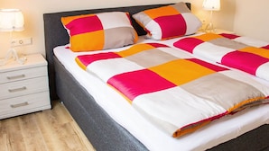 A neatly made bed with a striped bedspread in white, pink, and gray, flanked by two bedside lamps and a white nightstand with drawers. - Apartment in Oberkirch (Oberkirch)