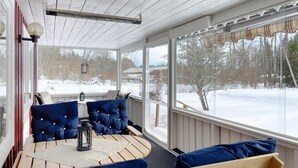 Une terrasse couverte avec une table en bois et des chaises rembourrées bleues, de grandes fenêtres offrant une vue sur un paysage enneigé.