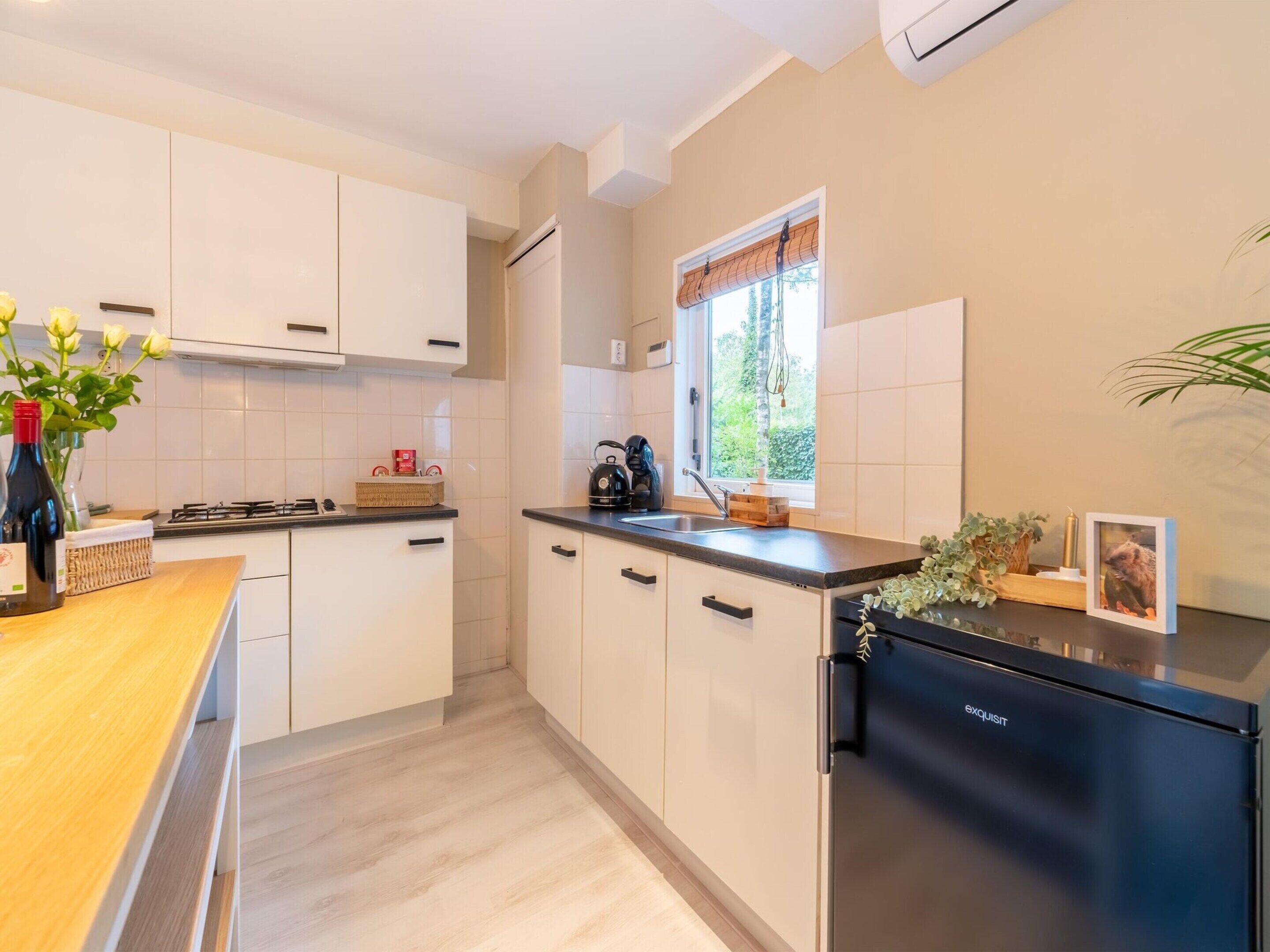 Une cuisine moderne avec des meubles blancs, un réfrigérateur noir et une fenêtre donnant sur un paysage verdoyant.
