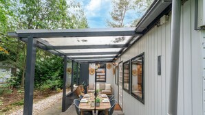Une terrasse couverte avec une table et des chaises, entourée d’un bâtiment blanc et de verdure.