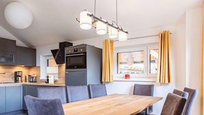 A modern kitchen with a central island, a dining area with a wooden table and upholstered chairs, and a window with curtains. - Amazing Chalet With Terrace (Stadl-Predlitz)