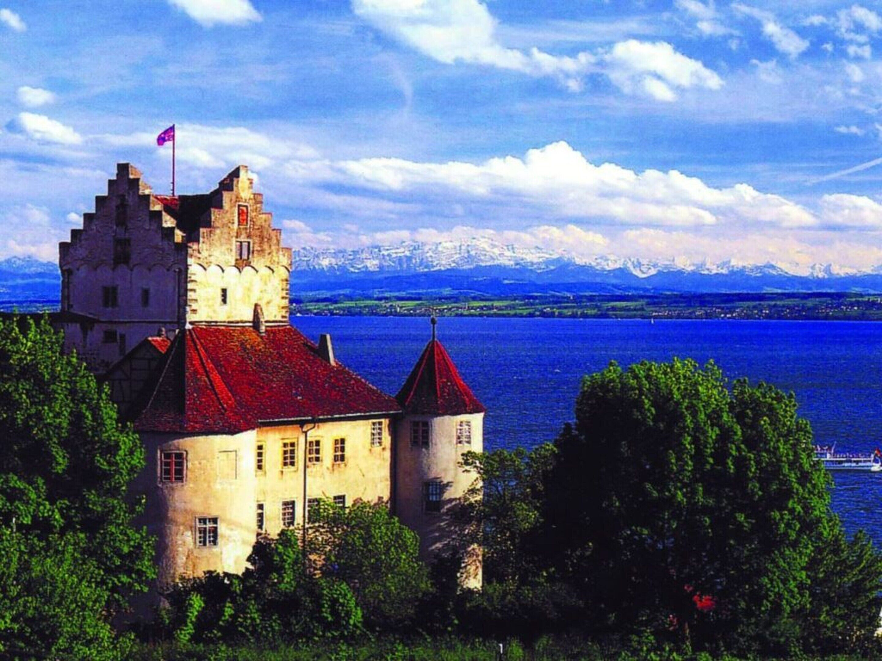A castle with a red roof, surrounded by trees and a lake with mountains in the background.