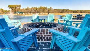 A poolside area with blue Adirondack chairs, a brick fire pit, and a view of a lake with trees in the background. - Resort style, beach town cottage, 5 mins from the beach- family friendly. (Beaufort)