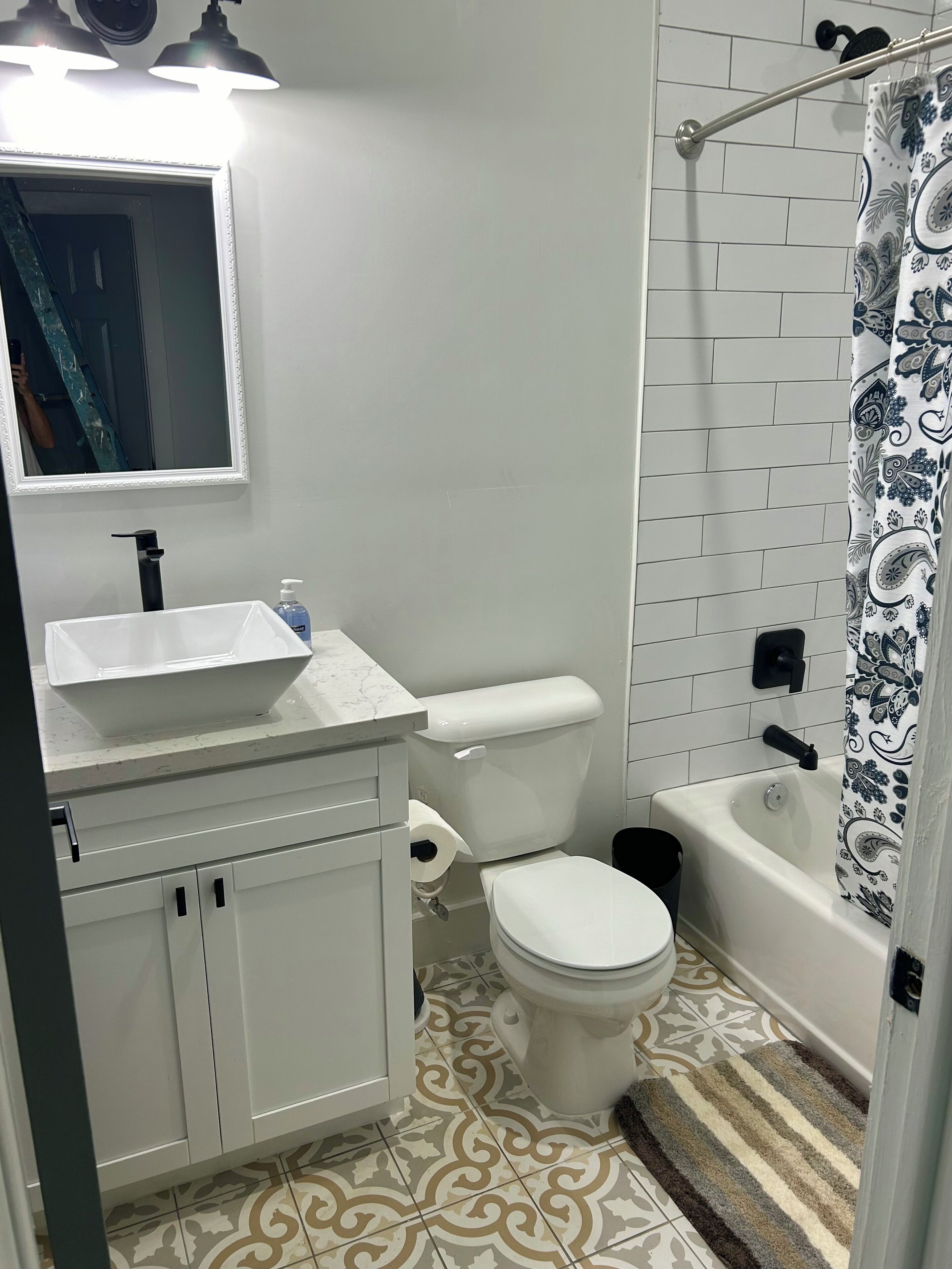 Une salle de bain avec un lavabo, des toilettes et une baignoire, tous de couleur blanche. Un miroir est placé au-dessus du lavabo, et il y a un rideau de douche orné d’un motif cachemire.