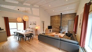 A spacious living area with a wooden floor, a large sofa, a dining table with chairs, and a kitchen area with a stone wall and a fireplace.
