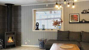 Un salon avec un poêle à bois, un canapé, une table basse, une fenêtre avec des stores et des objets décoratifs sur le rebord de la fenêtre.