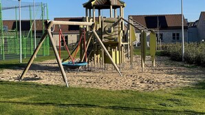 A playground with a swing set and climbing structures, surrounded by a fenced area and a building in the background. - Holiday Home in Koksijde Near Sint-andre Beach (Koksijde)