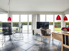 A spacious living area with a dining table, chairs, a TV, and large windows offering a view of the outdoors.
