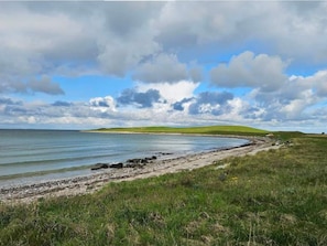 Ett kustlandskap med en gräsklädd strand, stilla vatten och böljande kullar i bakgrunden.